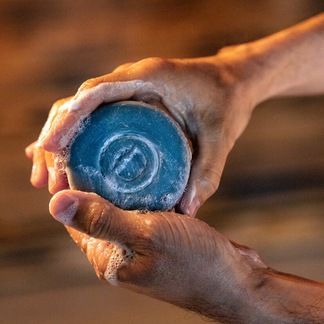 A person holding a Beardbrand Utility Bar in their hands. The bar is a charcoal gray color with a Beardbrand logo in the center and is covered in soap suds.