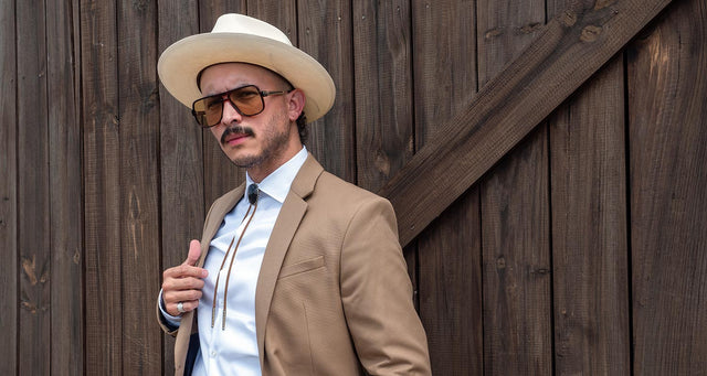 A man standing in front of a barn door and wearing a tan suit with a white collared shirt and bolo tie. 