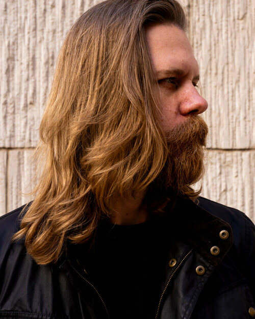 A man with long, wavy, golden-brown hair and a thick, full beard gazes off to the side. He is wearing a black jacket with brass buttons against a textured beige wall.